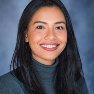 A light brown lady with black hair and brown eyes smiling for a corporate headshot with a deep blue background