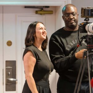 Smiling woman stands beside a man operating a professional video camera on a tripod indoors, both looking at the rig.